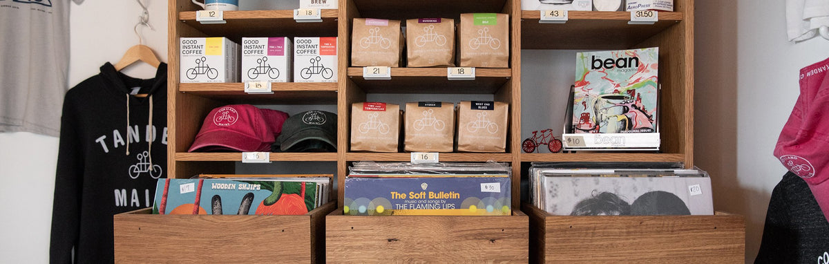 Wooden shelves displaying coffee bags, assorted hats, magazines, and records. Tandem-branded merchandise includes t-shirts, a sweatshirt, and a pink bandana. Price tags are visible on most items.