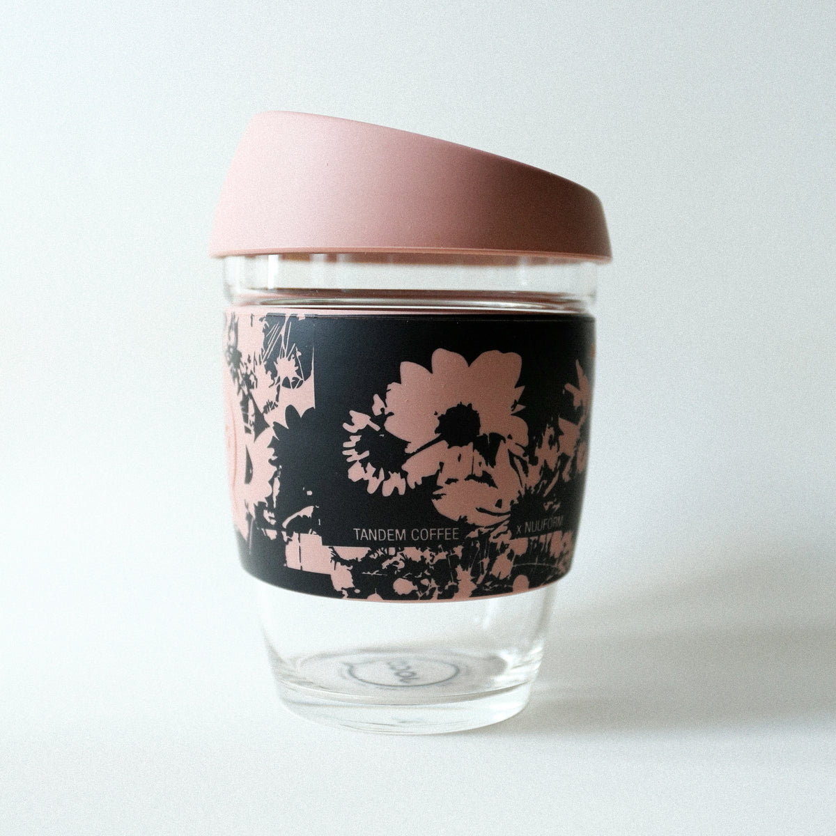 The Tandem x NUUFORM Joco Cup by Tandem Coffee Roasters is a plastic-free, reusable borosilicate glass coffee cup with a pink lid and a black sleeve decorated with pink floral patterns, shown on a white background.