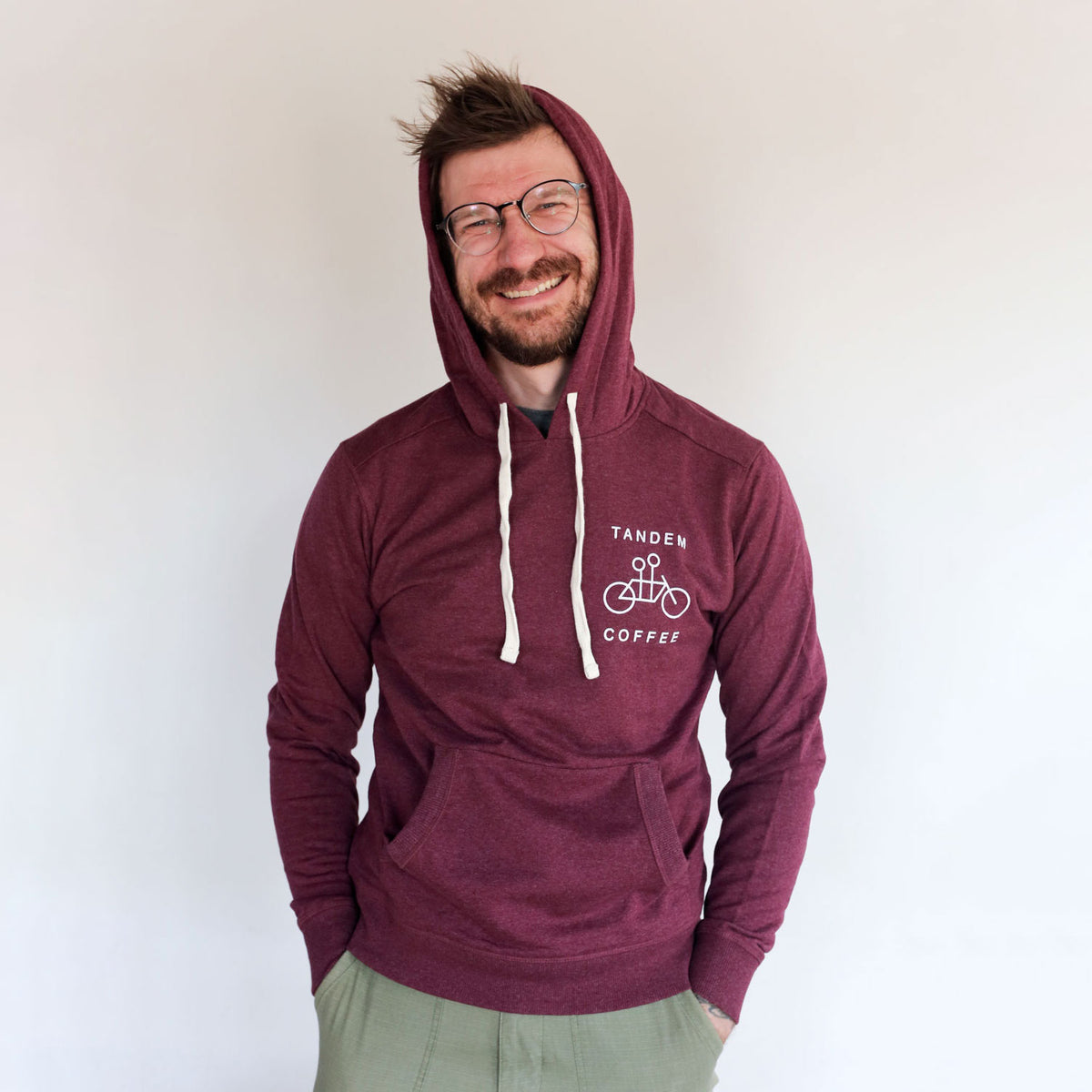 A cheerful man with glasses wearing a maroon Recycled Pullover Hoodie from Tandem Coffee Roasters, standing against a plain background, smiling broadly at the camera.