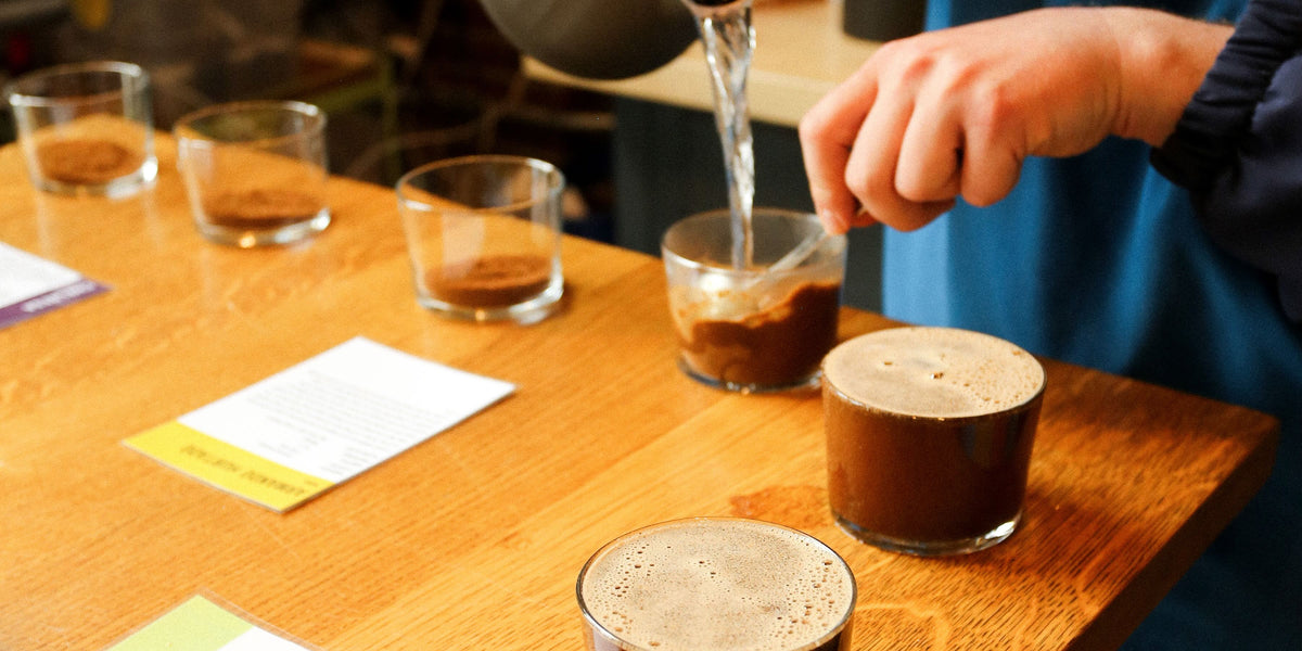 A close-up of a person pouring hot water from a kettle into a small glass containing ground coffee, set on a wooden table with other similar glasses.