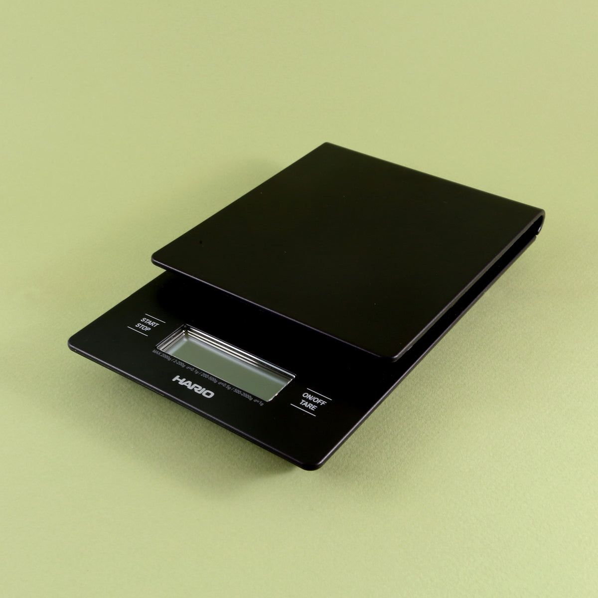 A black Hario V60 Drip Scale with a visible LCD display, sitting on a pale green background. The scale is compact and has a sleek, modern design, ideal as a home coffee scale.